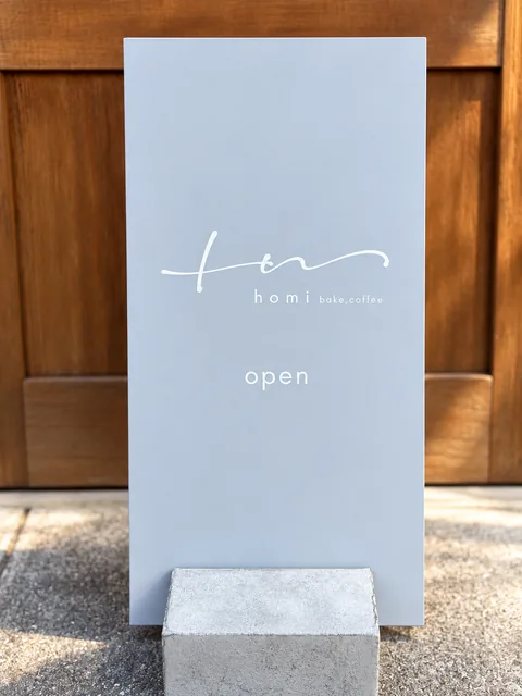 木の扉の前に置かれた、青みがかった灰色の立て看板。筆記体のロゴ、homi bake,coffee、open の文字